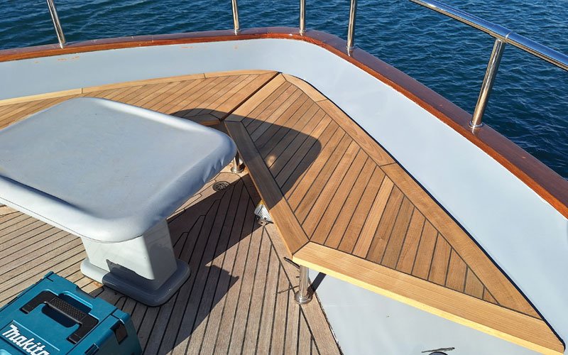 A close-up of a yacht's teak deck, featuring a raised, curved step with a symmetrical plank pattern. The deck has a warm, natural wood tone with visible grain and black caulking lines.
