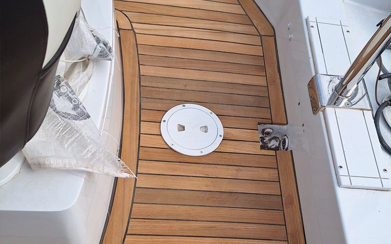 A close-up of a yacht's teak deck, featuring a raised, curved step with a symmetrical plank pattern. The deck has a warm, natural wood tone with visible grain and black caulking lines.