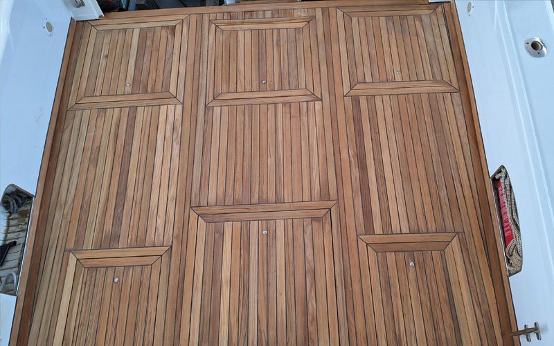 up of a yacht's teak deck, featuring a raised, curved step with a symmetrical plank pattern. The deck has a warm, natural wood tone with visible grain and black caulking lines.