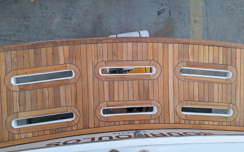 up of a yacht's teak deck, featuring a raised, curved step with a symmetrical plank pattern. The deck has a warm, natural wood tone with visible grain and black caulking lines.