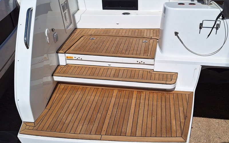 up of a yacht's teak deck, featuring a raised, curved step with a symmetrical plank pattern. The deck has a warm, natural wood tone with visible grain and black caulking lines.