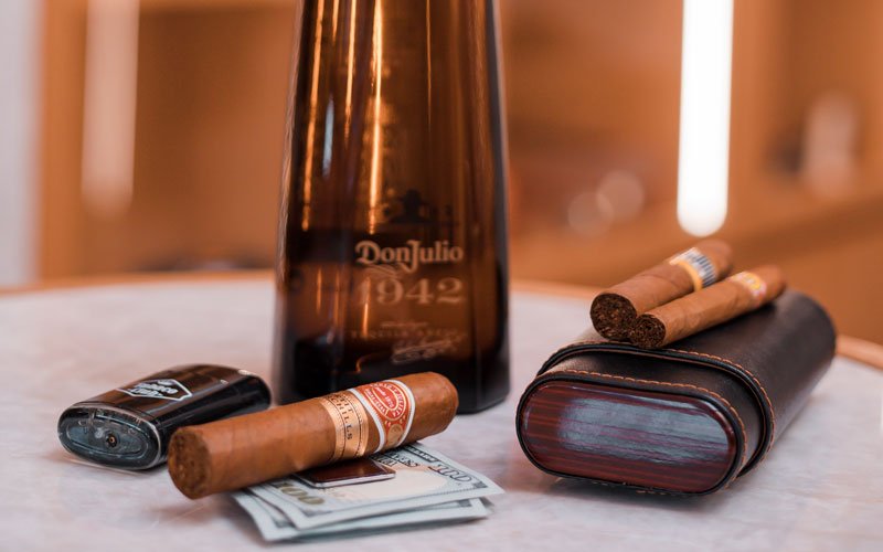 a bottle of Don Julio 1942 tequila on a table, surrounded by cigars, a cigar case, a lighter, and several US dollar bills