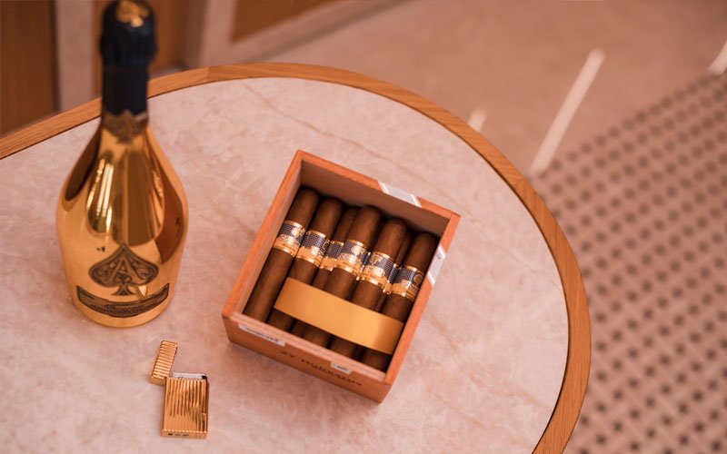a gold bottle of Armand de Brignac champagne, a gold lighter, and a wooden box of cigars with gold bands, placed on a round marble table