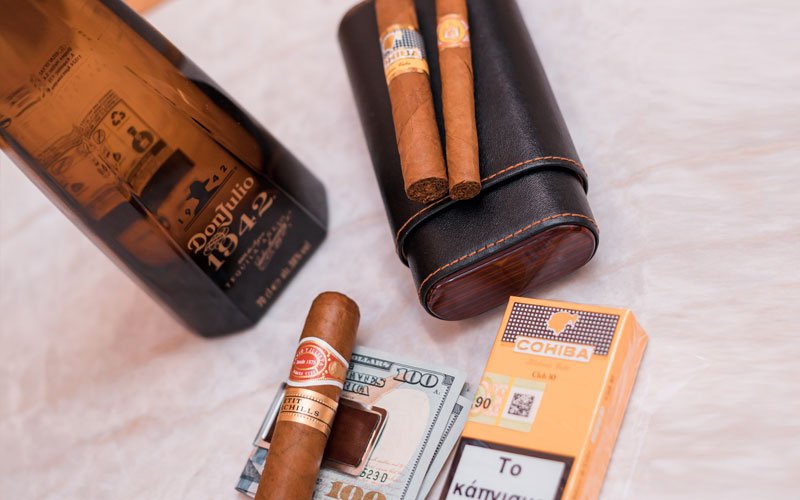 a bottle of Don Julio 1942 tequila on a table, surrounded by cigars, a cigar case, a lighter, and several US dollar bills