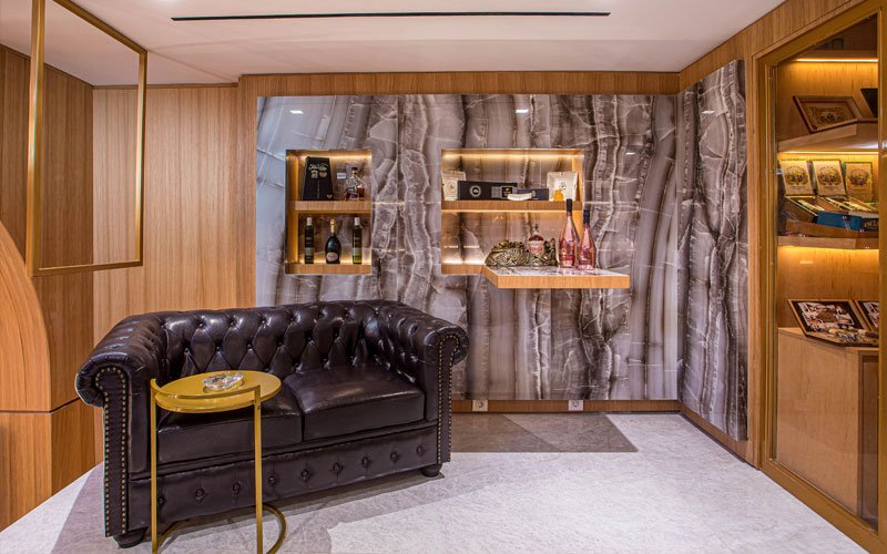 Luxurious cigar lounge corner featuring a dark leather Chesterfield sofa, a gold side table with an ashtray, backlit marble wall with shelves displaying cigars and premium bottles.