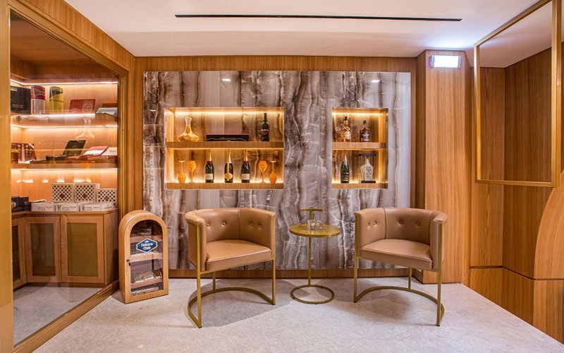 Elegant cigar lounge seating area with two modern leather armchairs, a small gold side table, illuminated marble wall shelves displaying spirits and glassware, and a humidor cabinet on the side.