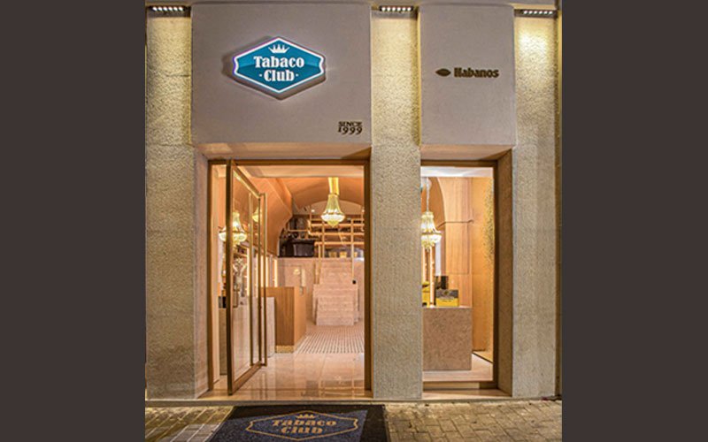 Exterior view of a stylish cigar shop entrance named 'Tabaco Club', with glass doors open, warm interior lighting, and a branded doormat in front. The facade includes a sign with the Tabaco Club logo and the Habanos brand
