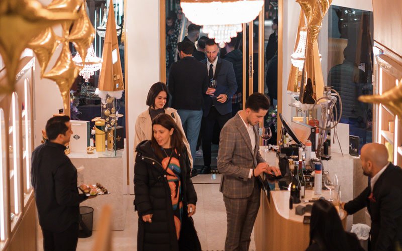 Interior view of a modern cigar lounge or luxury boutique during an event, with elegantly dressed people mingling, a bartender preparing drinks, and golden decorations with chandeliers hanging from the ceiling