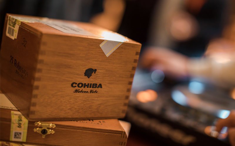 Close-up of a wooden cigar box labeled 'Cohiba Habana, Cuba' with a silhouette logo, placed on top of another similar box, with a blurred background of a person operating DJ equipment