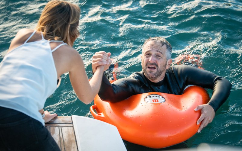 A man in the sea holding onto an orange lifebuoy, while a woman aboard a boat extends her hand to help him climb aboard