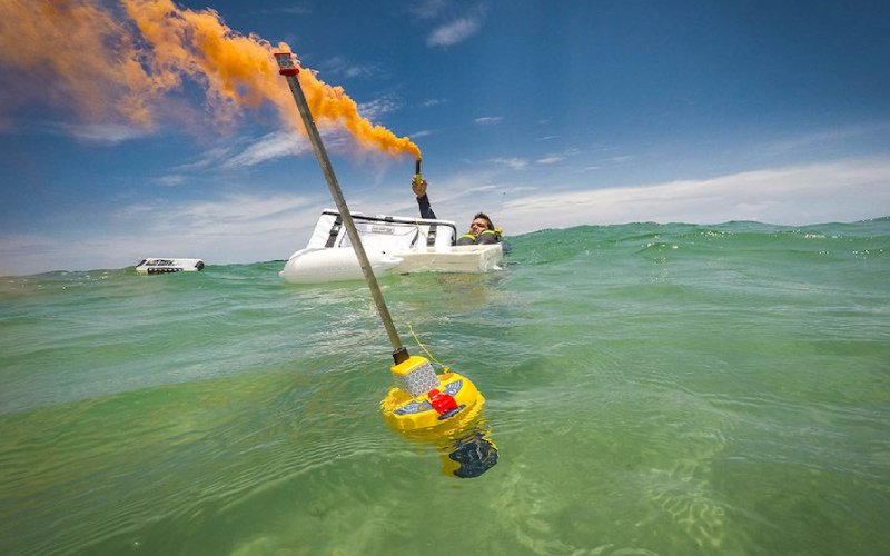 A person in a life raft at sea uses an orange smoke signal for rescue. In the foreground, a Versilia Marine Service emergency buoy with a pole and reflective tape floats on the water. The ocean is slightly rough, and another vessel is visible in the background under a clear blue sky