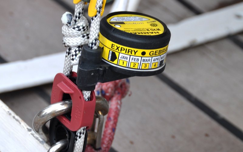 A close-up of a Versilia Marine Service safety release system attached to a rope with a metal shackle and red plastic clip. The black and yellow device has an expiry date indicator with months and numbers for tracking usage. The background shows part of a boat deck
