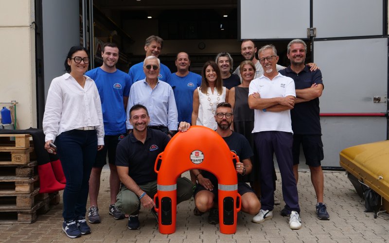 A group of employees from Versilia Marine Service pose in front of a workshop, smiling. Two team members kneel in the front, holding an orange U SAFE remote-controlled life-saving buoy. The background includes pallets, equipment, and an open workshop door