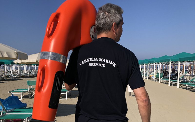 A staff member of Versilia Marine Service, seen from behind, wearing a black T-shirt with the company logo on the back, holding a U-SAFE lifebuoy while standing on an organized beach