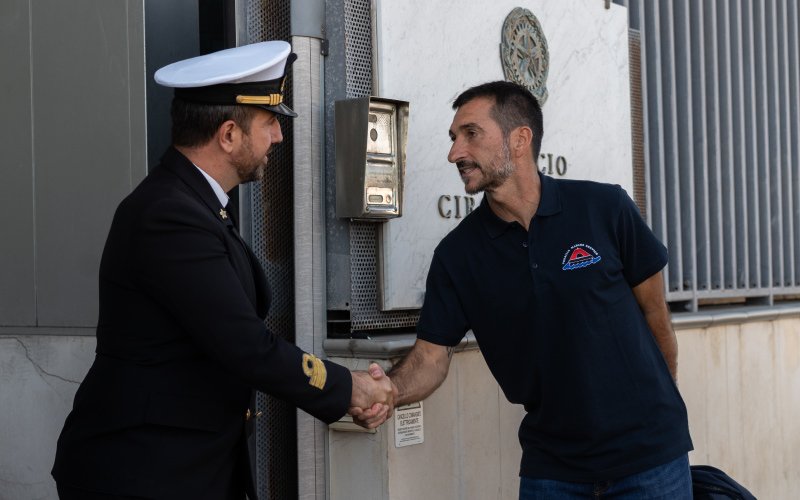 A man wearing a navy blue polo shirt with the Versilia Marine Service logo shakes hands with a uniformed officer outside a government or maritime office, as indicated by the official plaque on the wall behind them