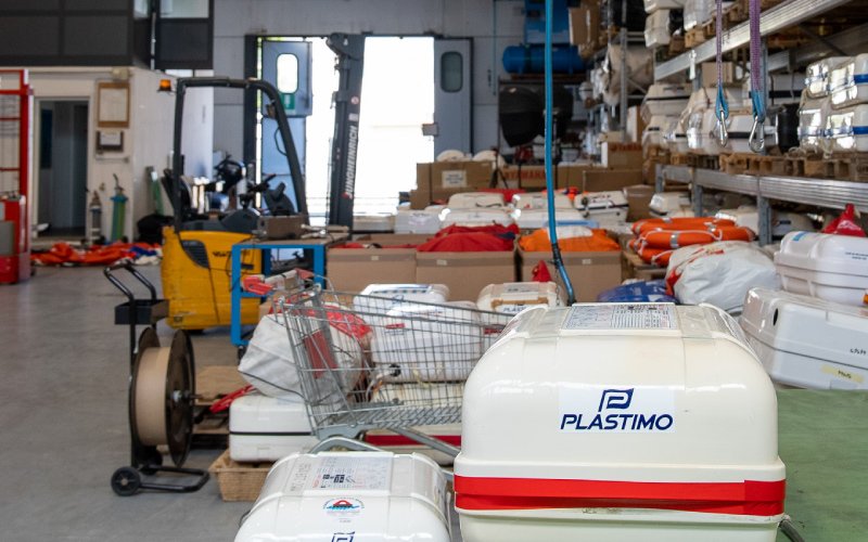 The Versilia Marine Service storage and maintenance facility, filled with marine safety equipment, including Plastimo life rafts, life jackets, and other survival gear. A forklift and metal shelves stocked with supplies are visible in the background
