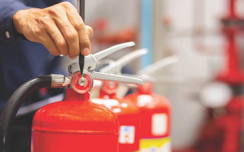 A close-up of a technician inspecting a red fire extinguisher, using a tool to check the pressure gauge. Several other fire extinguishers are lined up in the background, indicating a safety inspection or maintenance process