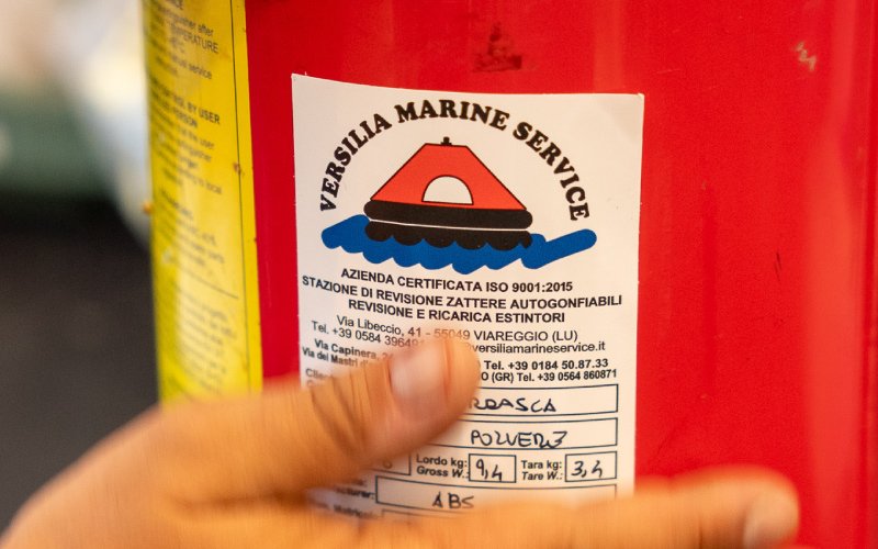 A close-up of a technician inspecting a red fire extinguisher, using a tool to check the pressure gauge. Several other fire extinguishers are lined up in the background, indicating a safety inspection or maintenance process