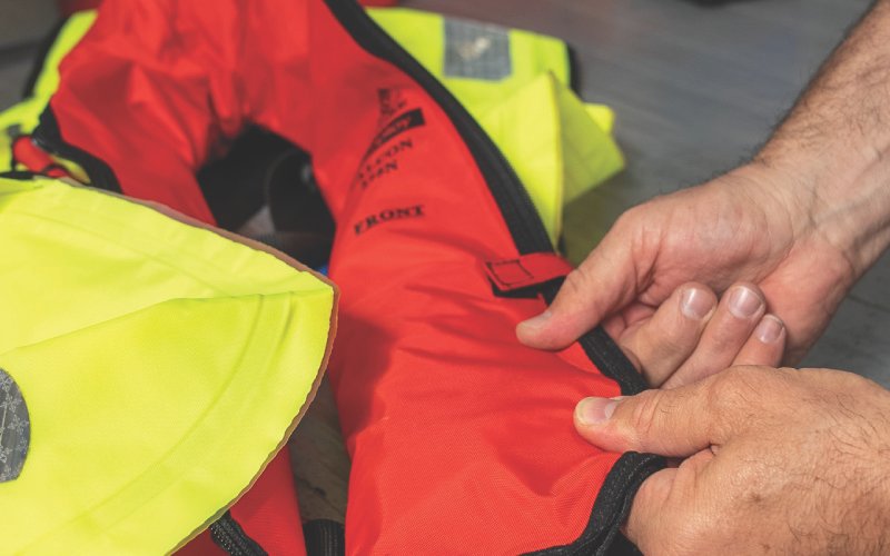 A close-up of a technician inspecting a bright red and yellow life jacket. The hands are carefully handling the flotation device, ensuring its condition and readiness for use. The life jacket features reflective patches and safety labels, indicating professional maintenance or quality control