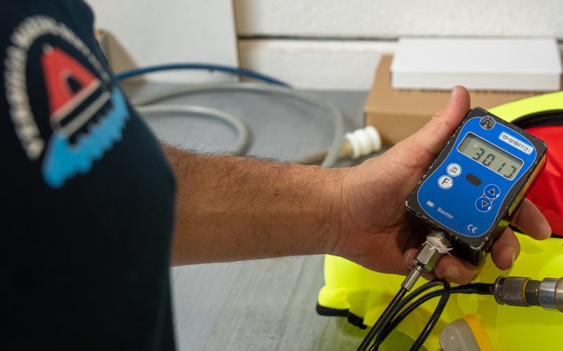 A technician wearing a navy-blue shirt with a maritime logo holds a digital pressure gauge displaying "30.17." The gauge is connected to a device, likely for testing the inflation pressure of a life-saving flotation device. In the background, a bright yellow and red life jacket is partially visible, along with coiled hoses and a workbench, indicating a professional marine safety inspection setting.