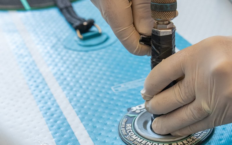 A close-up of a technician's gloved hands connecting an inflation or pressure-testing device to a valve on a blue inflatable surface.