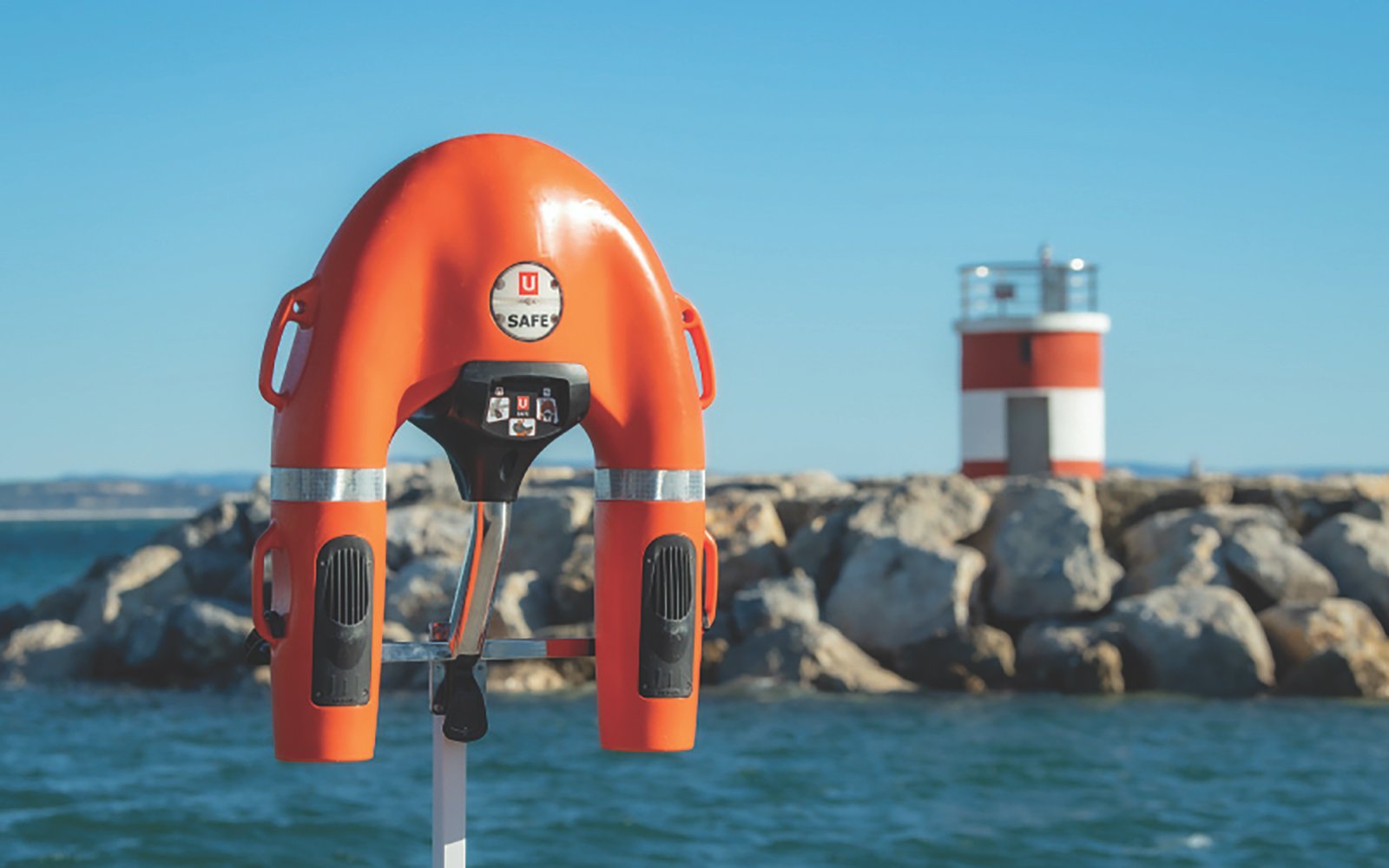 A U-shaped self-propelled lifebuoy with an orange body and black propulsion units mounted on a metal stand near the water. The device has the "U SAFE" logo on its center control panel, which features buttons for operation