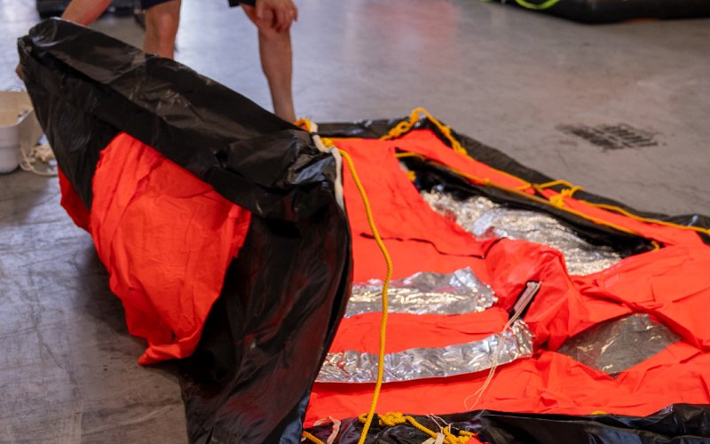 A partially inflated life raft with a bright orange and black exterior is being inspected indoors. The raft has reflective silver panels and yellow ropes along its edges. A person, partially visible in the background, is handling the raft