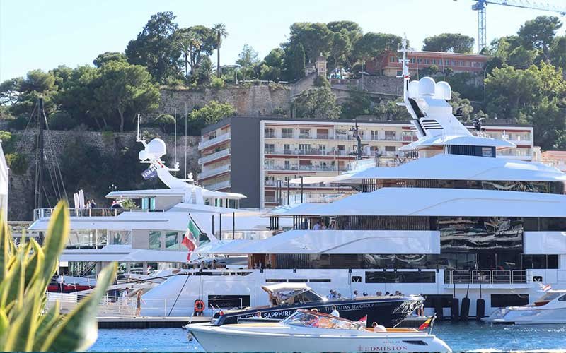 Luxury yachts docked in a marina with Italian flags, surrounded by speedboats and modern waterfront buildings, with a lush hillside and historic stone walls in the background.