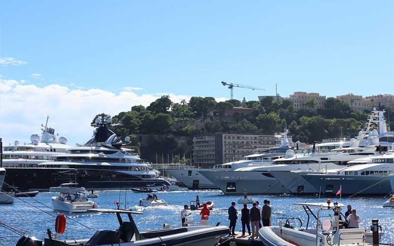 Luxury yachts docked in a marina with Italian flags, surrounded by speedboats and modern waterfront buildings, with a lush hillside and historic stone walls in the background.