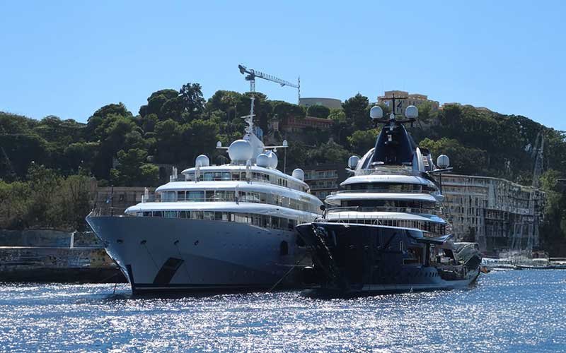 Two large luxury yachts docked side by side in a harbor with a forested hillside and buildings in the background under a clear blue sky
