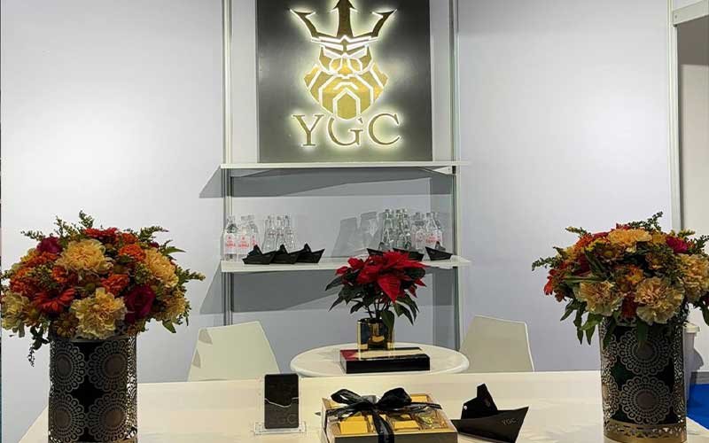 A modern exhibition booth or reception area featuring a glowing YGC logo on the wall, floral arrangements on both sides, a small round table with holiday-themed decor, and shelves with water bottles and glassware in the background