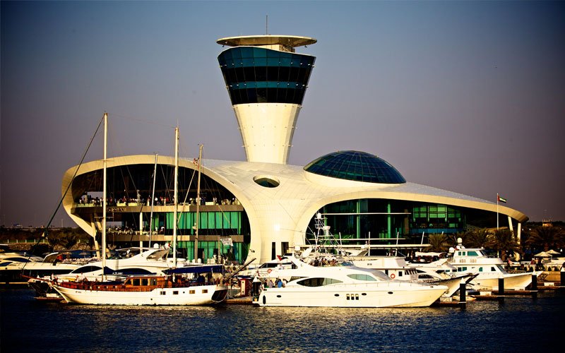 Yas Marina control tower and waterfront building illuminated at dusk with yachts in the foreground.
