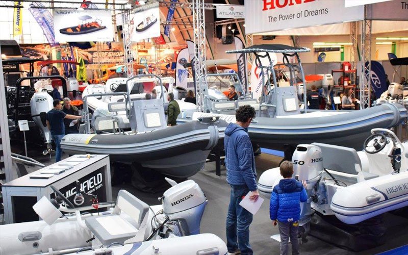 Indoor scene from the Zagreb Boat Show showcasing various inflatable boats and RIBs on display. The boats are arranged under bright exhibition lighting. Attendees are seen interacting with exhibitors, examining the boats and their features.