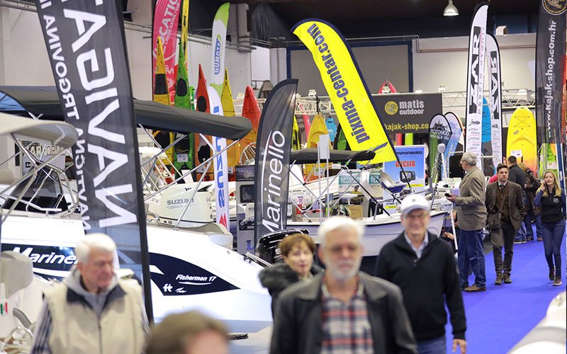 Indoor scene from the Zagreb Boat Show showcasing various inflatable boats and RIBs on display. The boats are arranged under bright exhibition lighting. Attendees are seen interacting with exhibitors, examining the boats and their features.