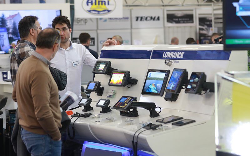 Indoor scene from the Zagreb Boat Show showcasing various monitors and electrical systems. Attendees are seen interacting with exhibitors, examining the equipment and their features.