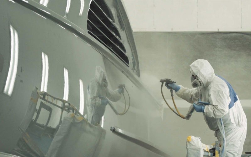 A technician wearing a protective suit and specialized mask is spray-painting a yacht using a spray gun, focusing on precision and even coverage